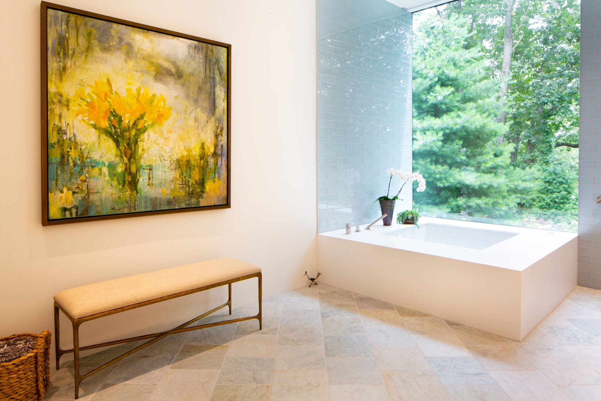 A photo of a bathroom with a large white square tub with light blue tiles on its walls and a ceiling-height window. To the left is a bench, towel basket, and a large framed painting of yellow flowers.