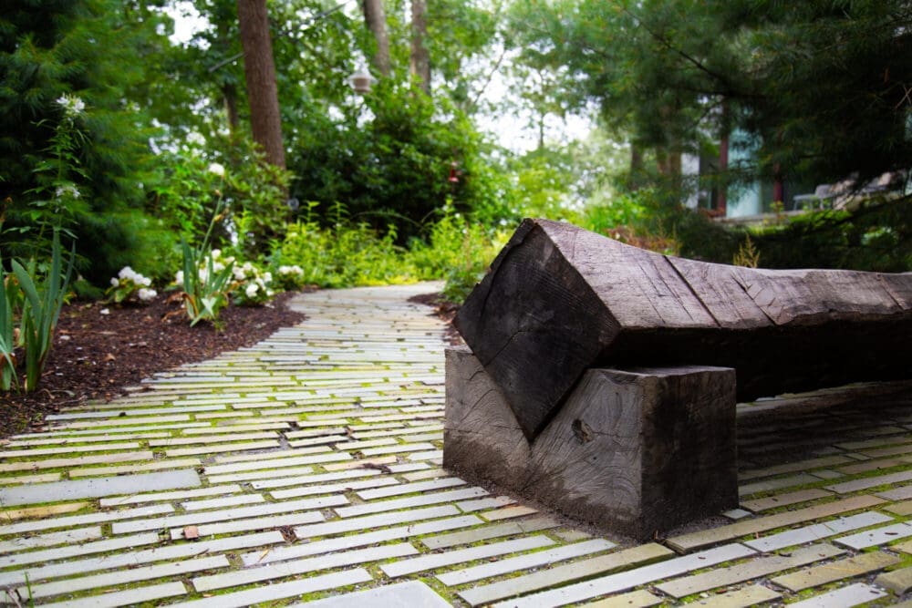 A photo of a diamond-shaped wooden bench, a stone path, and greenery.