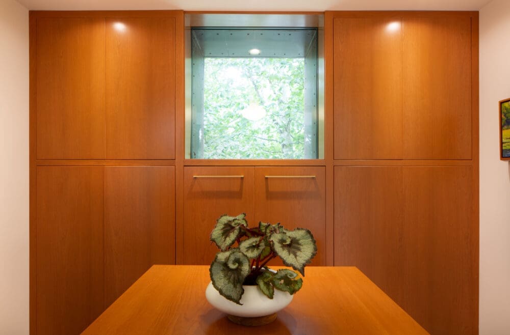 A photo of a walk-in closet made of cherry wood with a rectangular window. A large storage island and painted-leaf begonia is in the middle of the room.