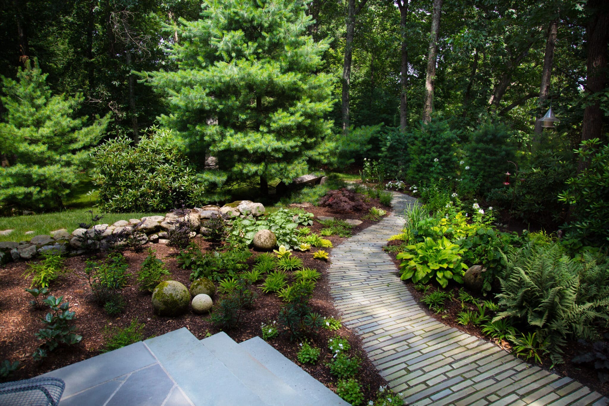 A photo of a stone pathway with a landscaped garden and coniferous trees on either side.