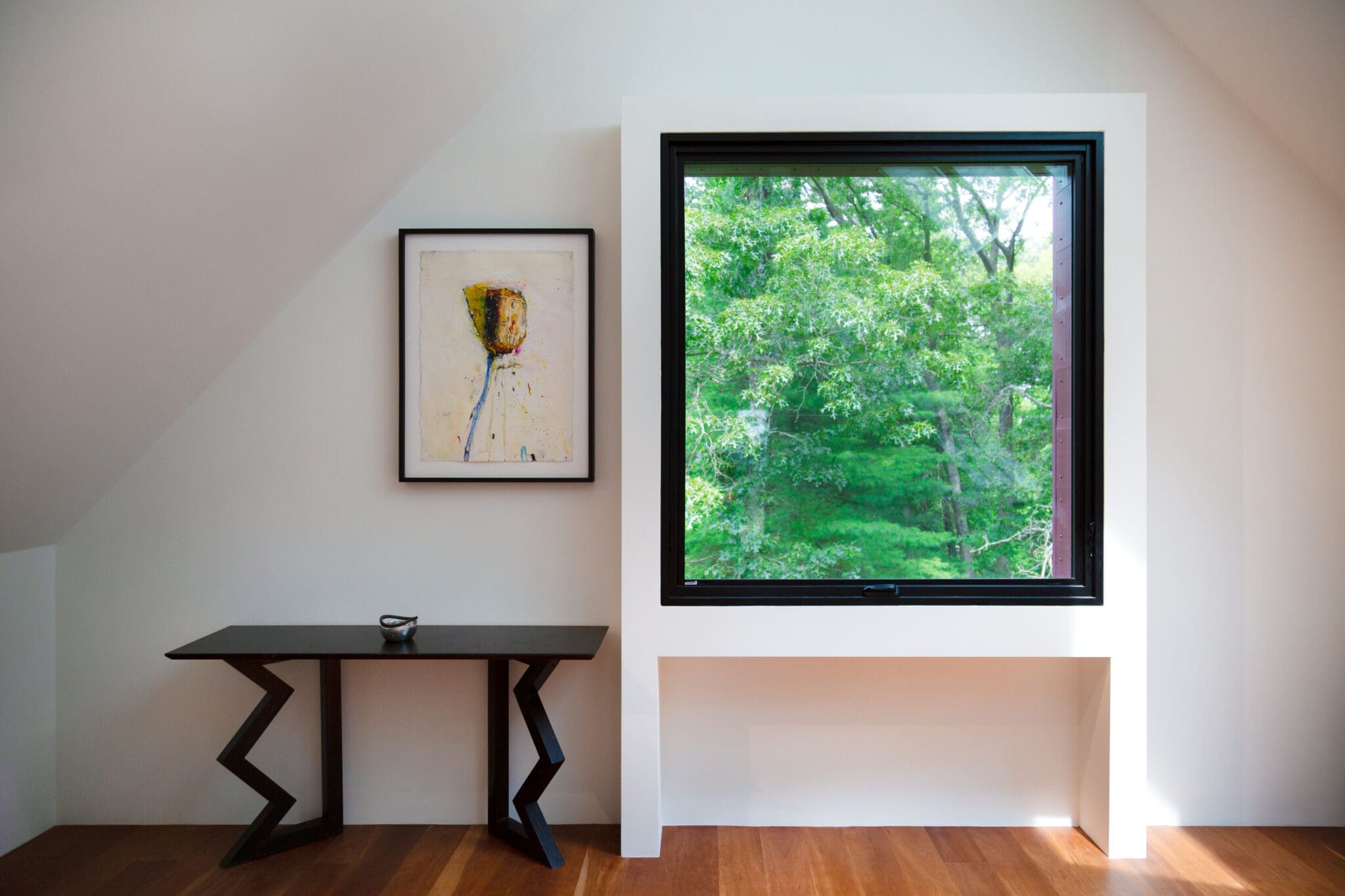 A photo of a room with vaulted ceilings, hardwood flooring, a large square window, a painting of a flower, and a modern black table.