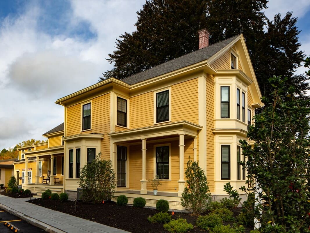 A yellow house with a porch and trees in the background.